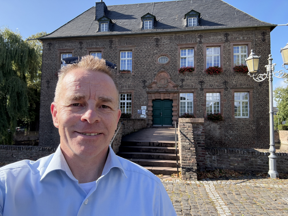 Mann in Hemd lächelt vor einem historischen Gebäude mit Ziegelfassade und grüner Tür. Sonniges Wetter, Laterne rechts im Bild. - Udo Diepmann, BarmeniaGothaer Merzenich