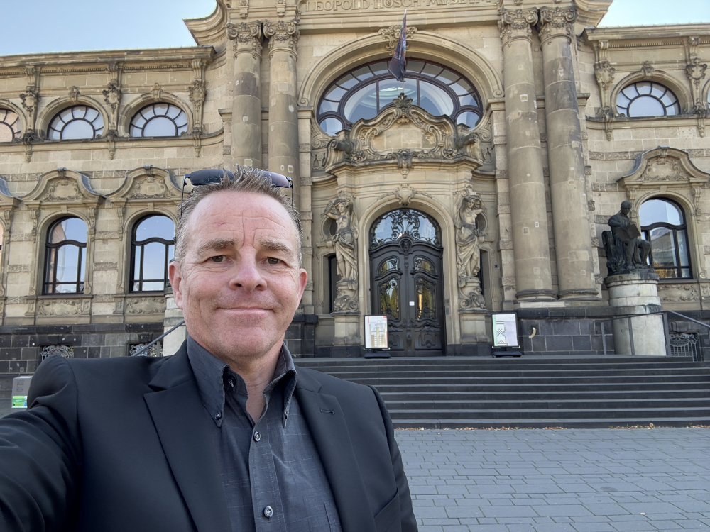 Ein Mann in einem schwarzen Anzug steht vor einem historischen Gebäude mit kunstvollen Steinsäulen und großen Fenstern. - Udo Diepmann, BarmeniaGothaer Merzenich