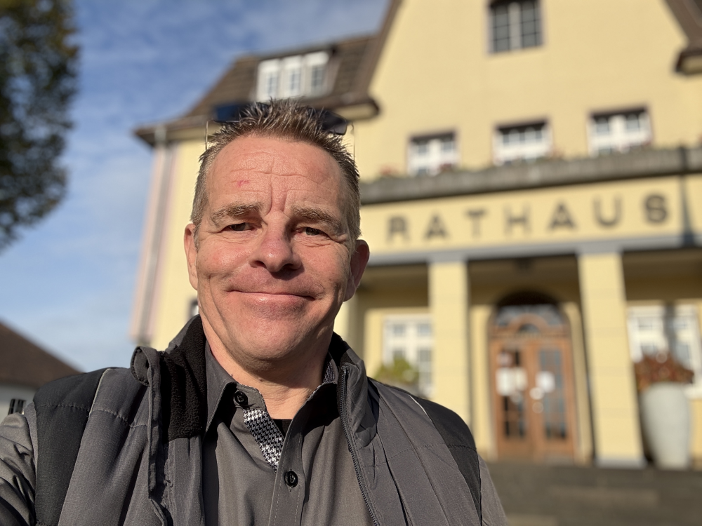 Ein Mann lächelt in die Kamera und steht vor einem Gebäude mit der Aufschrift "Rathaus" im Sonnenschein. - Udo Diepmann, BarmeniaGothaer Merzenich