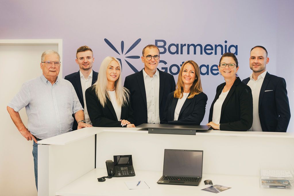 Sechs Personen stehen hinter einem Empfangstresen mit einem Laptop, vor einer Wand mit dem Logo von Barmenia Gothaer. - Andreas Klink, BarmeniaGothaer Bad Liebenzell