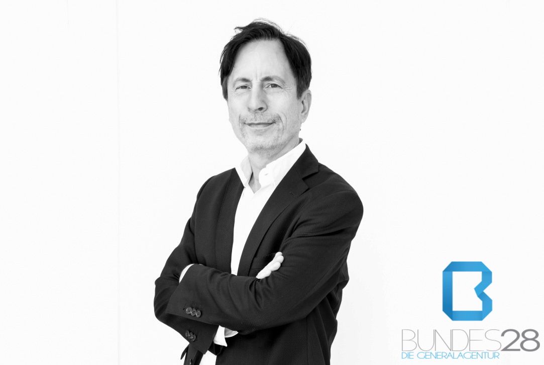 Ein Mann in Anzug mit verschränkten Armen lächelt leicht. Im Hintergrund ist das Logo "BUNDES28, Die Generalagentur" sichtbar. - Christian M. Schuchert, BarmeniaGothaer Berlin