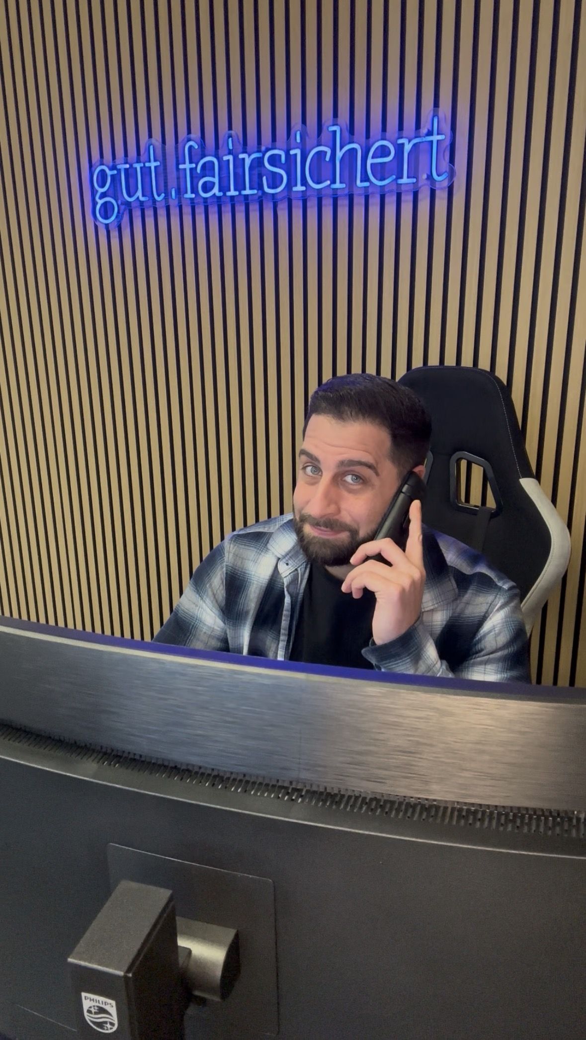 Ein Mann sitzt vor einem Computer und telefoniert. Über ihm leuchtet ein Schild mit der Aufschrift "gut.fairsichert". Im Hintergrund Holzverkleidung.