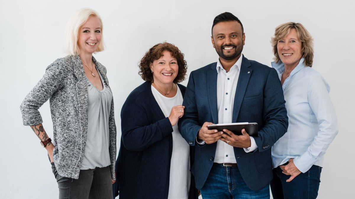 Vier lächelnde Personen stehen nebeneinander. Der Mann in der Mitte hält ein Tablet. Alle tragen Freizeit- oder Businesskleidung. - Thuvaragan Nesapalan e.K., BarmeniaGothaer Remscheid