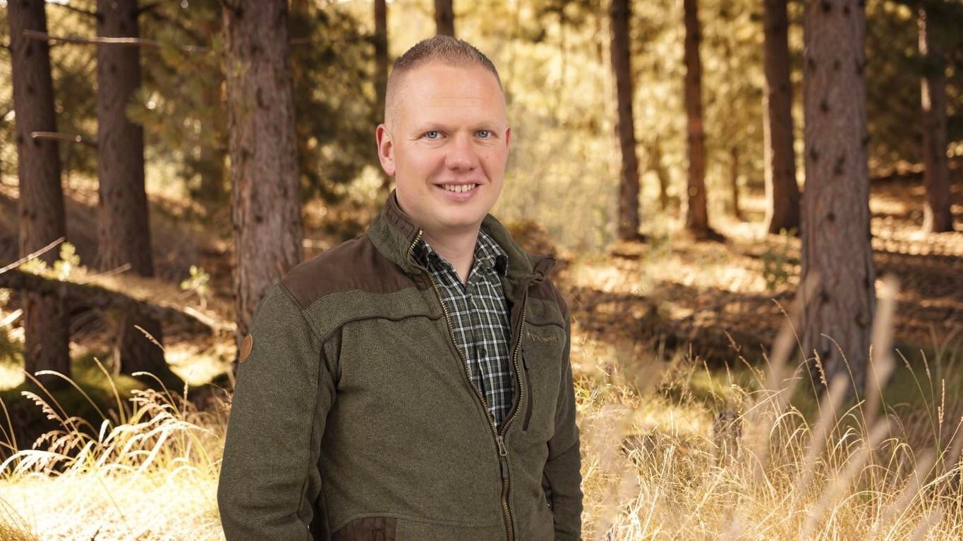 Ein Mann steht lächelnd in einem lichterfüllten Wald, trägt eine grüne Jacke und ein kariertes Hemd. Im Hintergrund sind hohe Bäume zu sehen.