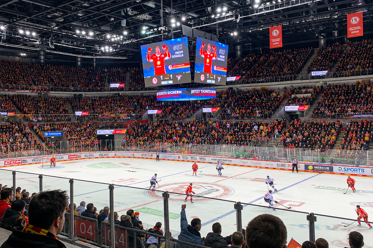 Ein vollbesetztes Stadion bei einem Eishockeyspiel, ein großes Display zeigt Informationen über einen Spieler, während Teams auf dem Eis spielen.