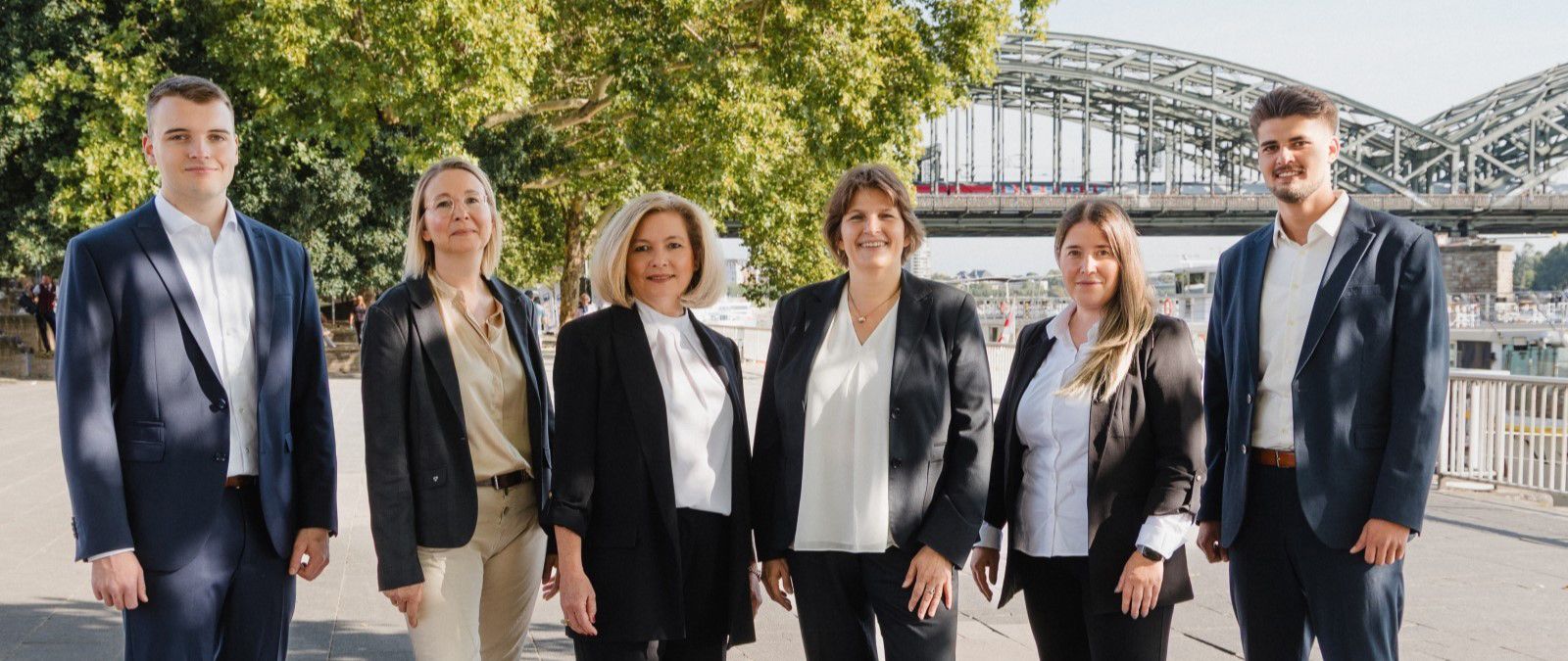 Sechs Personen in Businesskleidung stehen vor einem Baum und einer Eisenbahnbrücke und lächeln in die Kamera. - Martina Baedorf, BarmeniaGothaer Köln-Poll