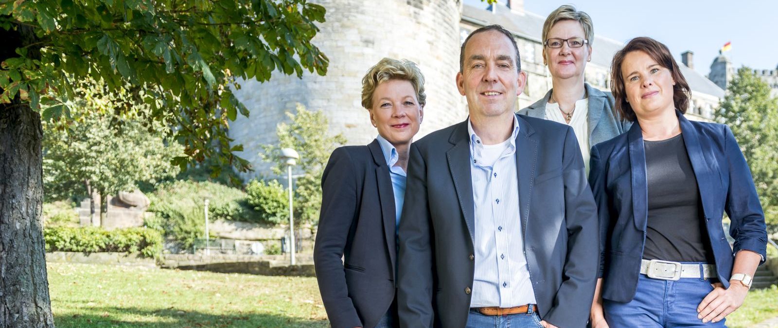Vier Personen in Geschäftsanzügen stehen im Freien vor einem Gebäude und lächeln in die Kamera. Bäume und blauem Himmel im Hintergrund. - Henkjan Morsink, BarmeniaGothaer Bad Bentheim