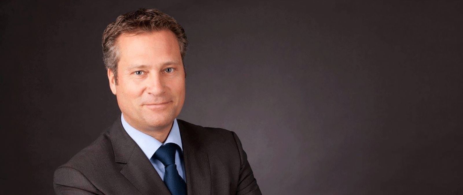Ein Mann im Anzug mit blauem Hemd und Krawatte, lächelnd vor einem dunklen Hintergrund. - Reiner Felden, BarmeniaGothaer Aachen