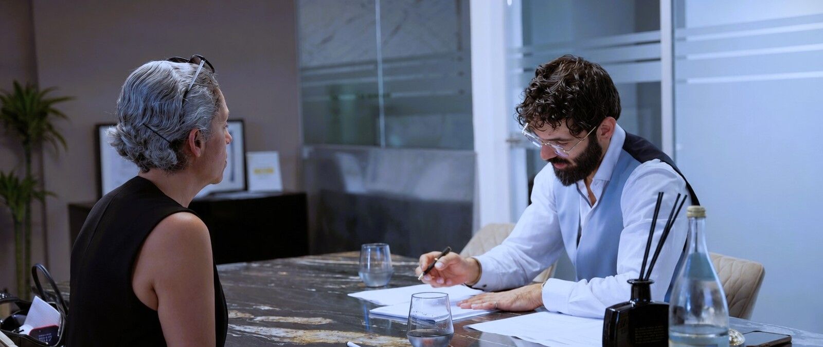 Zwei Personen in einem Büro im Gespräch, eine Frau mit grauen Haaren und ein Mann mit Bart, beide sitzen an einem Tisch mit Gläsern und Dokumenten. - Serhat Ocak, BarmeniaGothaer Düsseldorf