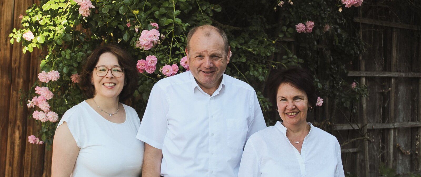 Drei lächelnde Personen in weißen Hemden stehen vor einem blühenden Rosenstrauch in einem Garten. - Ludwig Kandler, BarmeniaGothaer Offenberg