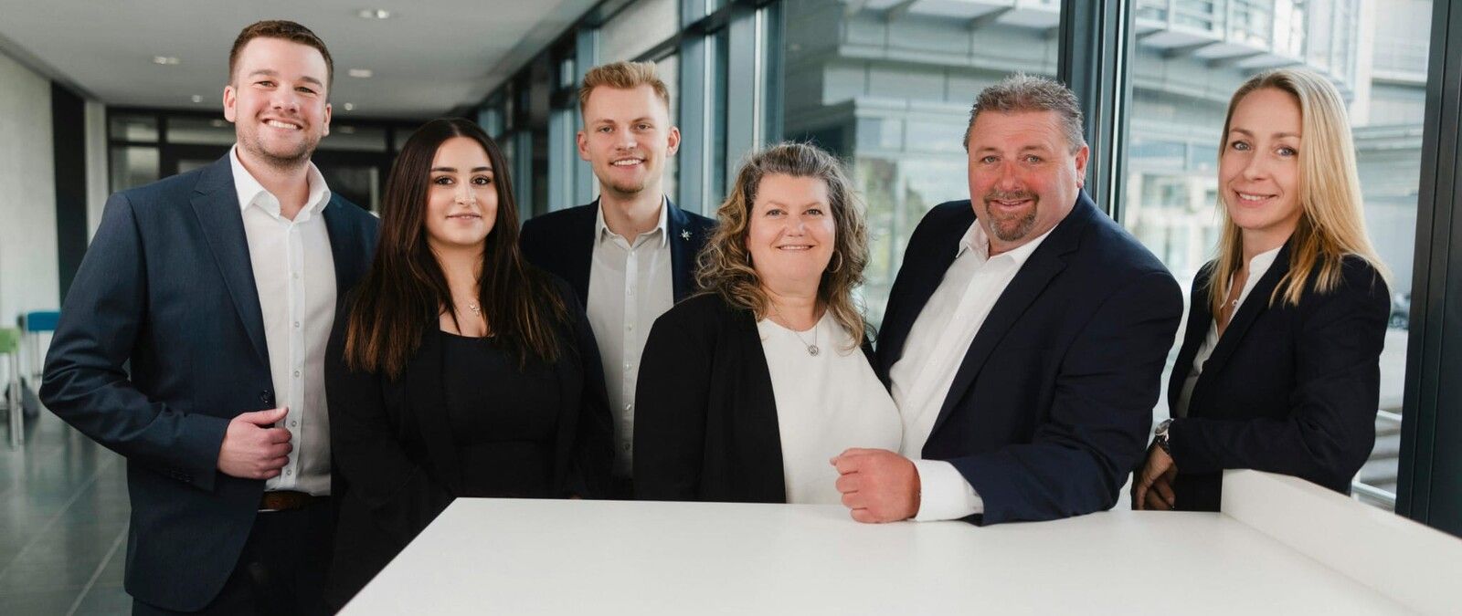 Sechs Personen in formeller Geschäftskleidung posieren lächelnd in einem modernen Bürogebäude mit Glaswänden. - Karsten Rostock, BarmeniaGothaer Frechen-Königsdorf