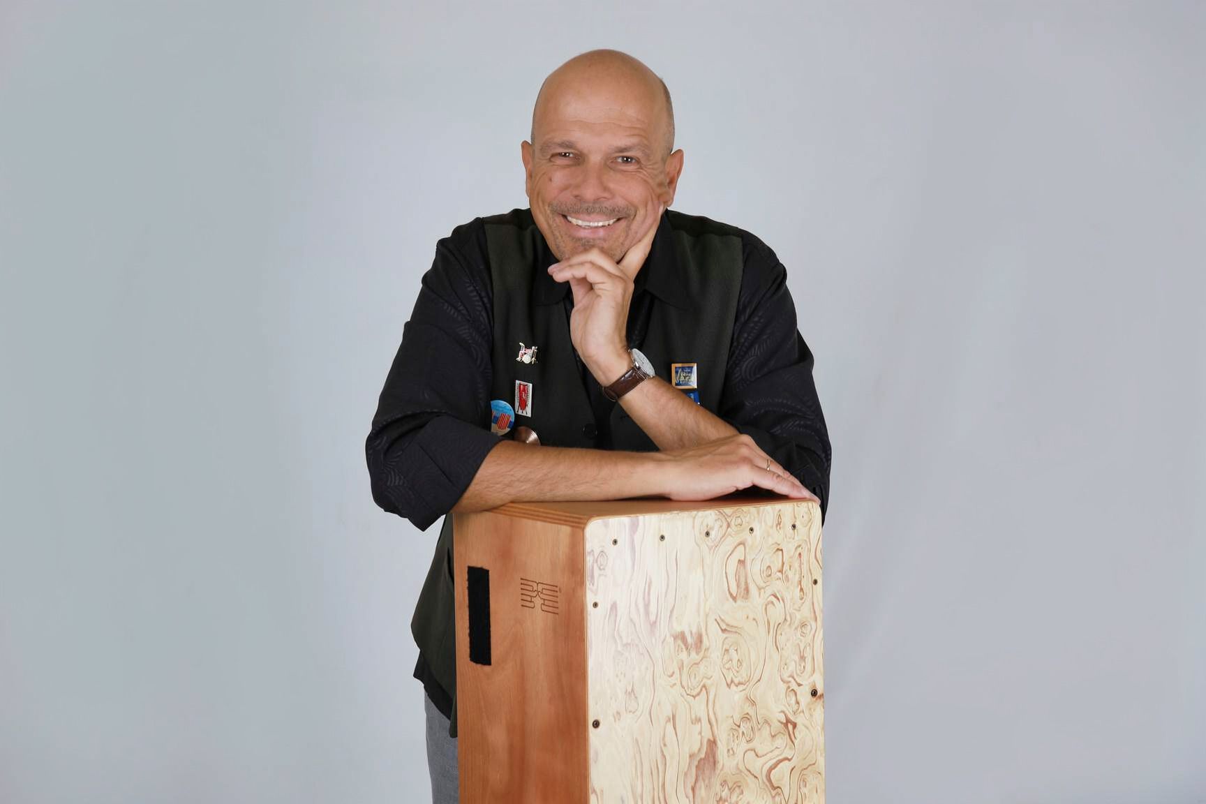 Ein lächelnder Mann lehnt sich auf eine Cajón, trägt eine schwarze Weste mit Ansteckern und hat die Hand ans Kinn gelegt.