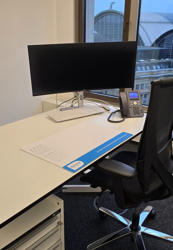 Moderner Schreibtisch mit Computer, Tastatur, Telefon und Kalenderunterlage. Großes Fenster mit Blick auf ein Gebäude mit gewölbtem Dach. - Kai-Erik Adler, BarmeniaGothaer Lollar