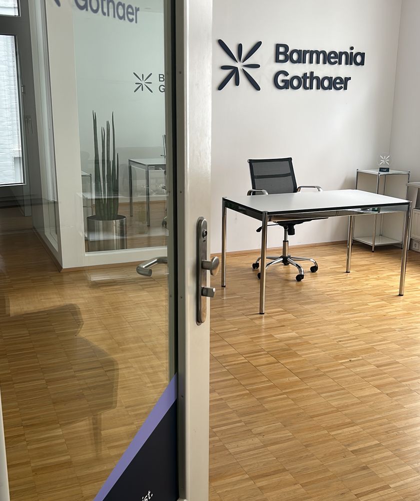 Ein Büro mit einem Schreibtisch, einem Stuhl und einem Wandlogo von "Barmenia Gothaer"; helle Holzböden sind sichtbar. - Chiara Dykgers, BarmeniaGothaer Wuppertal