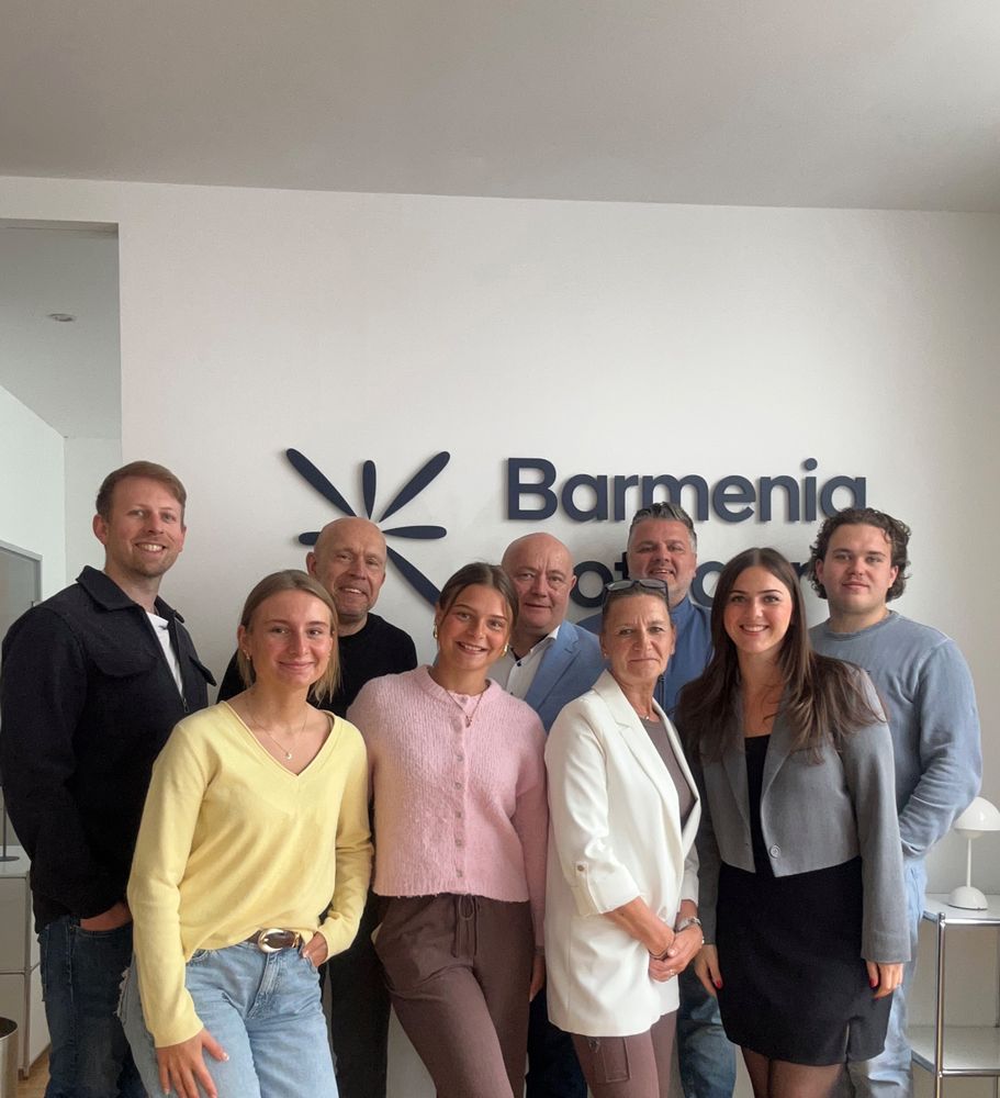 Gruppe von neun Menschen steht lächelnd vor einem Schild mit der Aufschrift "Barmenia" in einem Büro. - Chiara Dykgers, BarmeniaGothaer Wuppertal