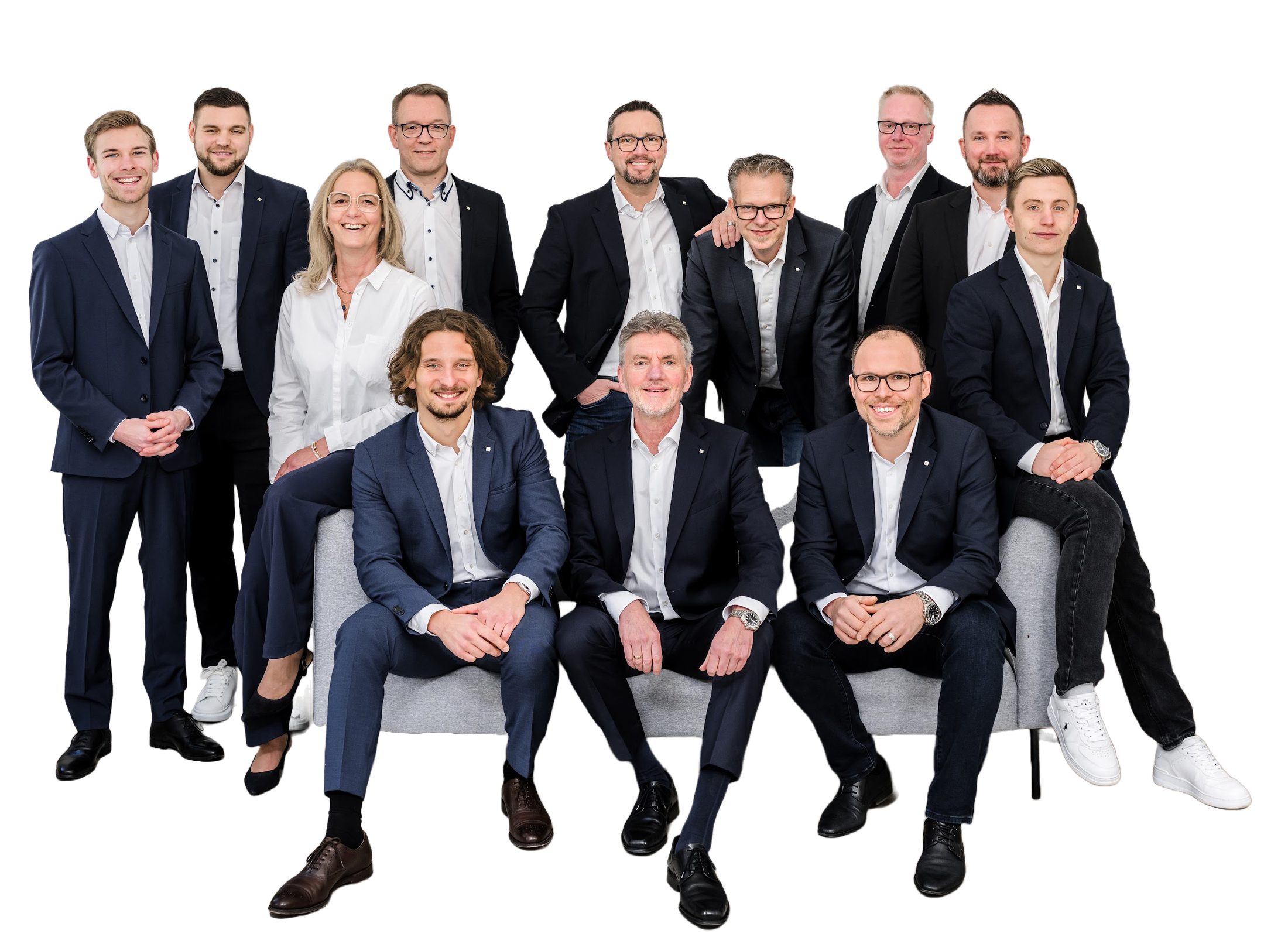 Gruppe von zwölf Männern und einer Frau in formeller Kleidung posieren zusammen im Studio, teils stehend und teils sitzend auf einem Sofa.