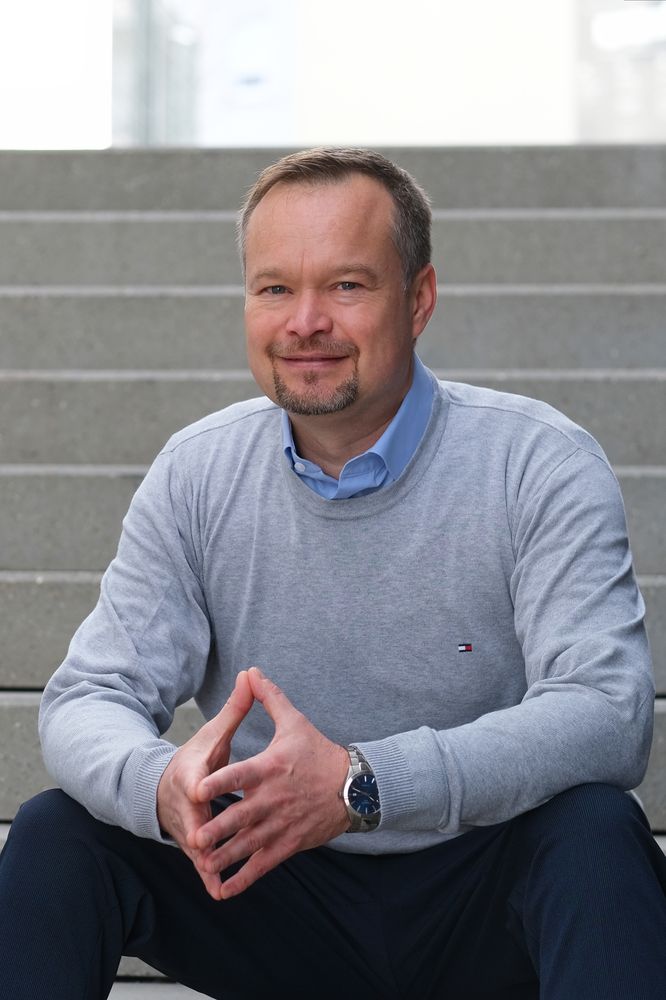 Ein Mann sitzt lächelnd auf einer Treppe, trägt einen grauen Pullover und eine blaue Uhr. - Tobias Bölling, BarmeniaGothaer Winnenden