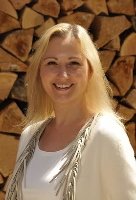Eine blonde Frau lächelt im Freien vor einem Hintergrund aus gestapeltem Brennholz. Sie trägt ein weißes Oberteil mit Fransen. - Hanno Wallrafen, BarmeniaGothaer Steinfurt