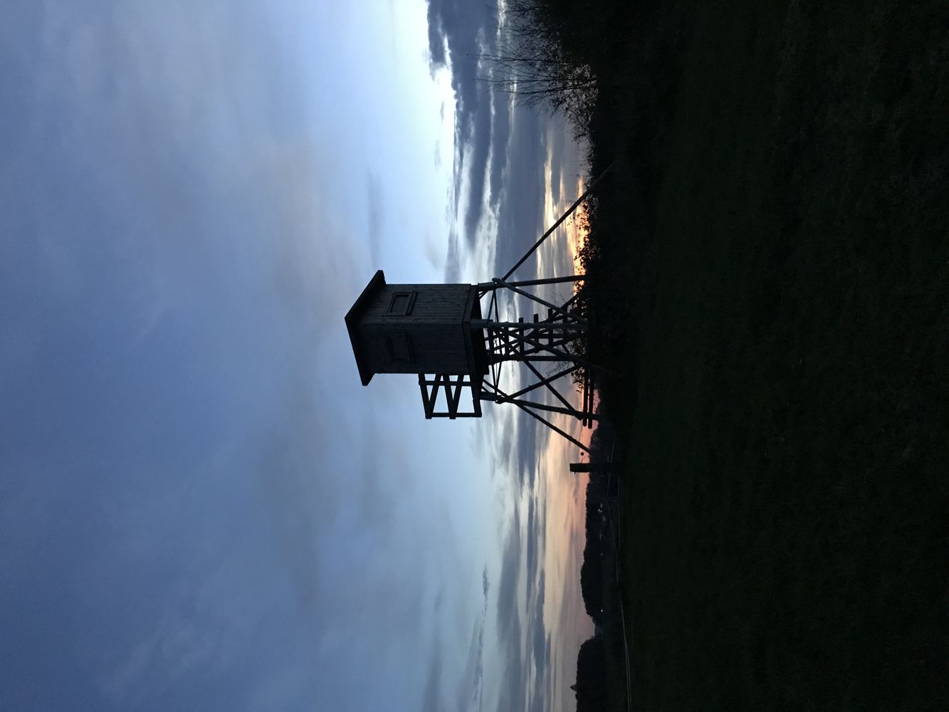 Silhouette eines Hochsitzes bei Sonnenuntergang, umgeben von Wiesen und Bäumen vor einem blau-grauen Himmel. - Sebastian Rolfs, BarmeniaGothaer Wipperfürth