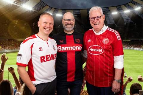 Drei Männer in Fußballtrikots verschiedener Vereine lächeln in einem beleuchteten Stadion in die Kamera.