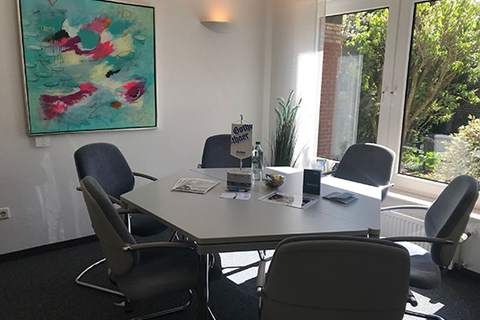 Heller Besprechungsraum mit fünfeckigem Tisch, fünf Stühlen, moderner Kunst an der Wand und großen Fenstern mit Blick auf einen grünen Garten.