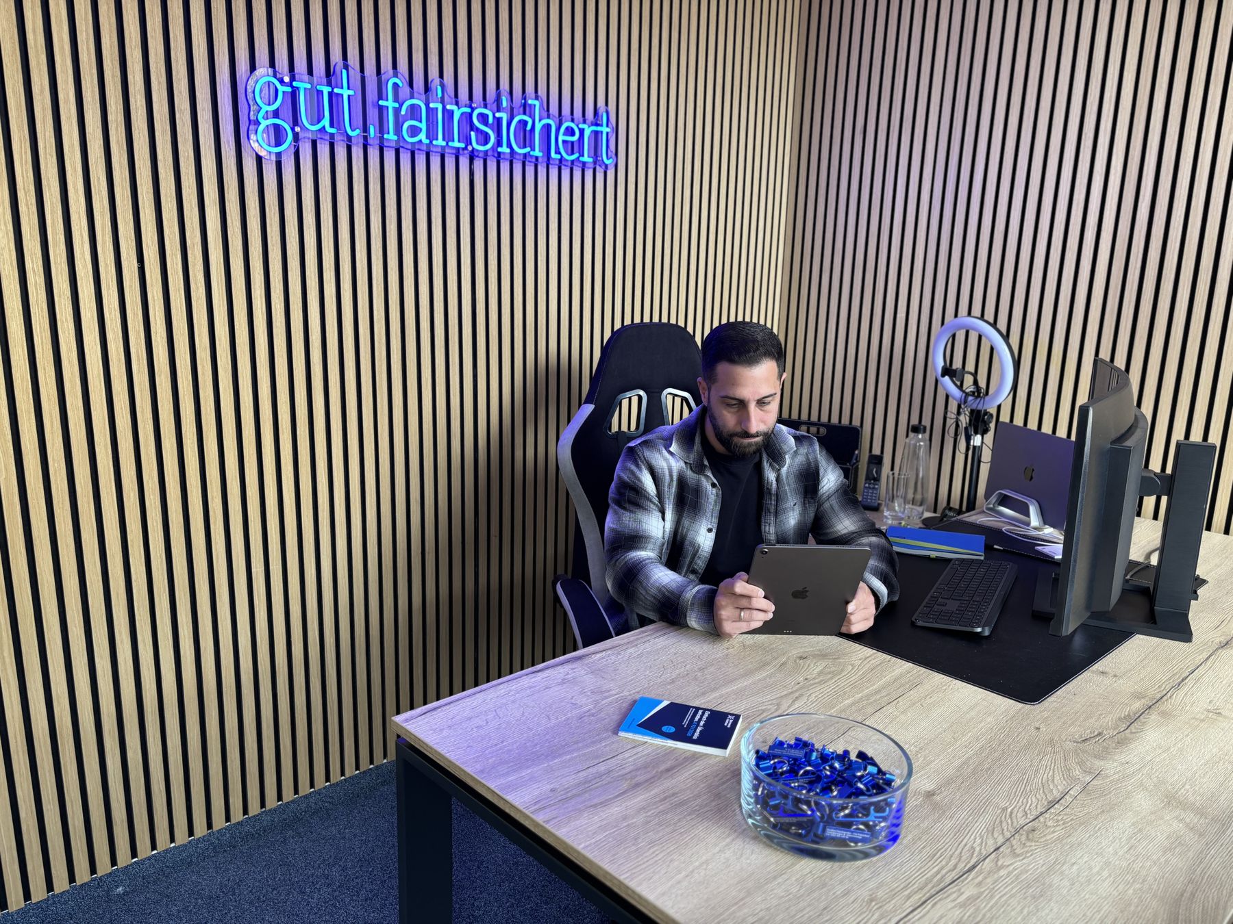 Mann mit Tablet sitzt an einem Schreibtisch in einem modernen Büro mit Holzverkleidung und LED-Schild "gut.fairsichert".