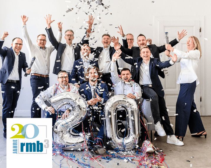 Gruppe von Menschen in Anzügen feiert mit Konfetti und Ballons. Ein Schild zeigt "20 Jahre rmb³".