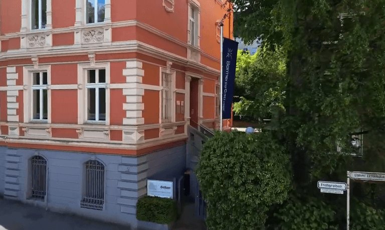 Rotes, historisches Gebäude an einer Straßenecke mit Beschilderung und grünem Laub an der Seite.