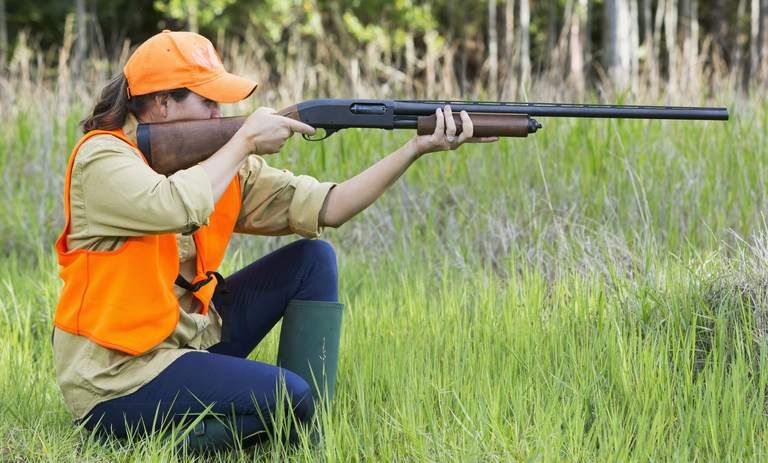 Jägerin in oranger Weste und Kappe kniet im Gras und zielt mit Gewehr in einem Waldgebiet.
