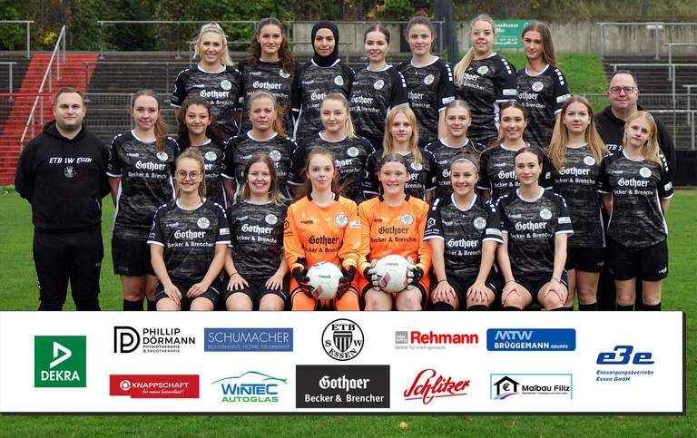 Eine Frauenfußballmannschaft posiert auf einem Fußballplatz. Alle Spielerinnen tragen einheitliche Trikots. Vorne sind Sponsorenlogos zu sehen.