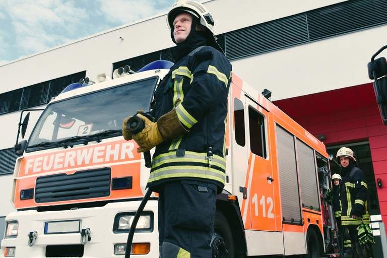 Ein Feuerwehrmann in Uniform steht vor einem Feuerwehrauto, bereit für den Einsatz. Ein Kollege ist im Hintergrund zu sehen.