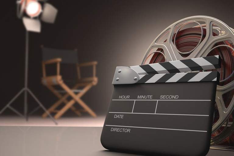 Filmklappe vor Filmrolle, Regiestuhl und Studiolicht im Hintergrund, die eine Filmszene andeuten.