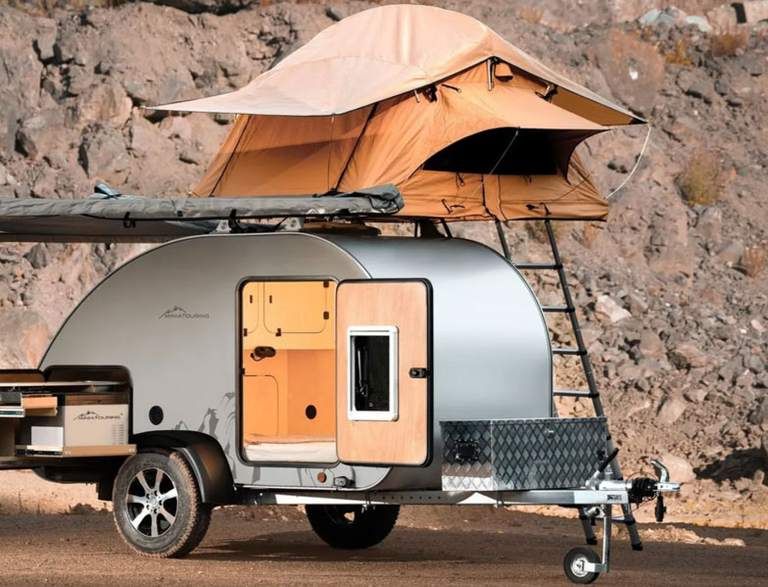 Silberner Campinganhänger mit ausgeklapptem Dachzelt und seitlicher Markise, vor einer felsigen Landschaft geparkt.
