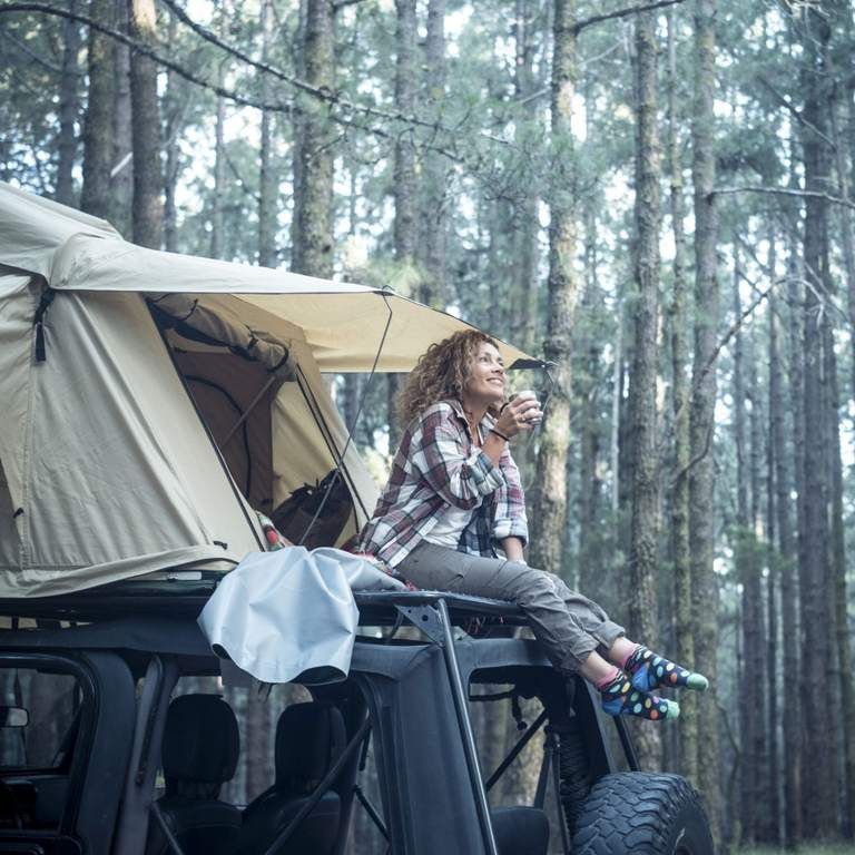 Frau sitzt auf Autodach mit Zelt in Wald und hält eine Tasse, lächelnd, in entspannter Camping-Atmosphäre.