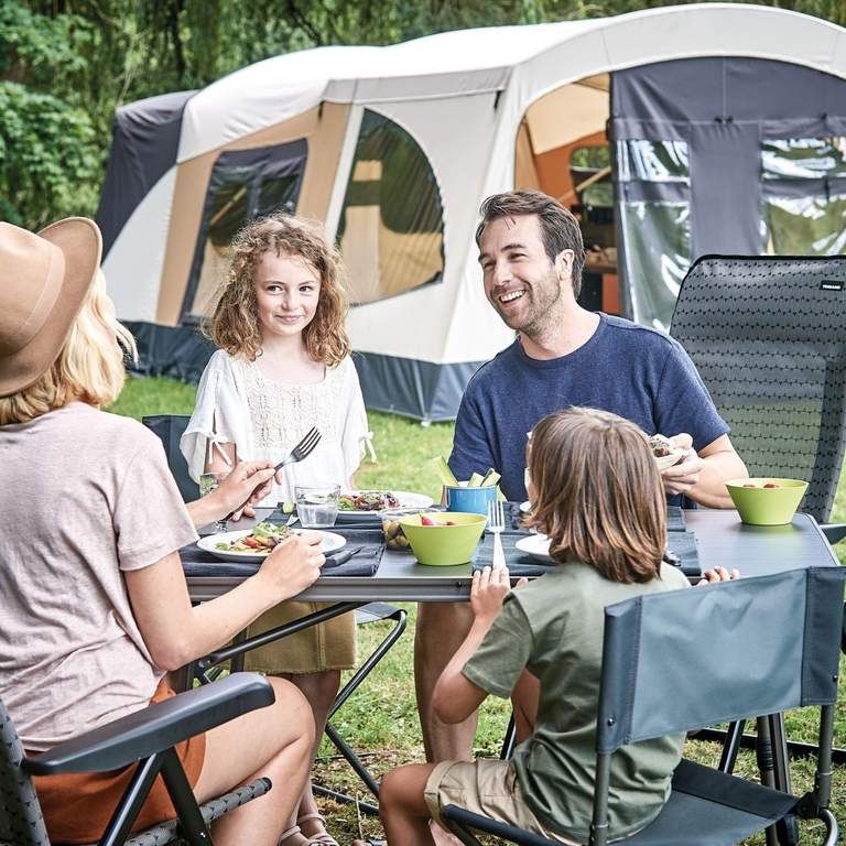 Eine Familie sitzt lachend beim Essen an einem Tisch im Freien vor einem großen Zelt.