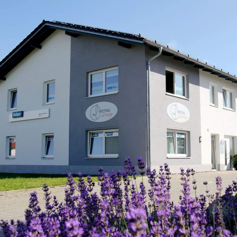 Zweistöckiges Gebäude mit grauer und weißer Fassade, im Vordergrund blühender Lavendel. Schilder mit "Meeting" an den Fenstern.