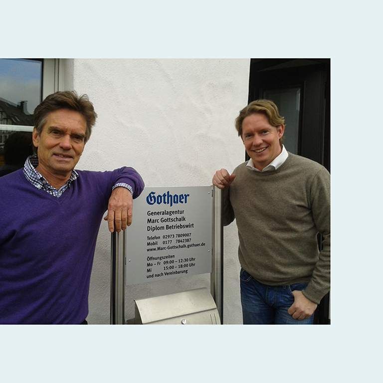 Zwei Männer stehen neben einem Schild der Gothaer Generalagentur Marc Gottschalk, das Kontaktinformationen und Öffnungszeiten anzeigt.