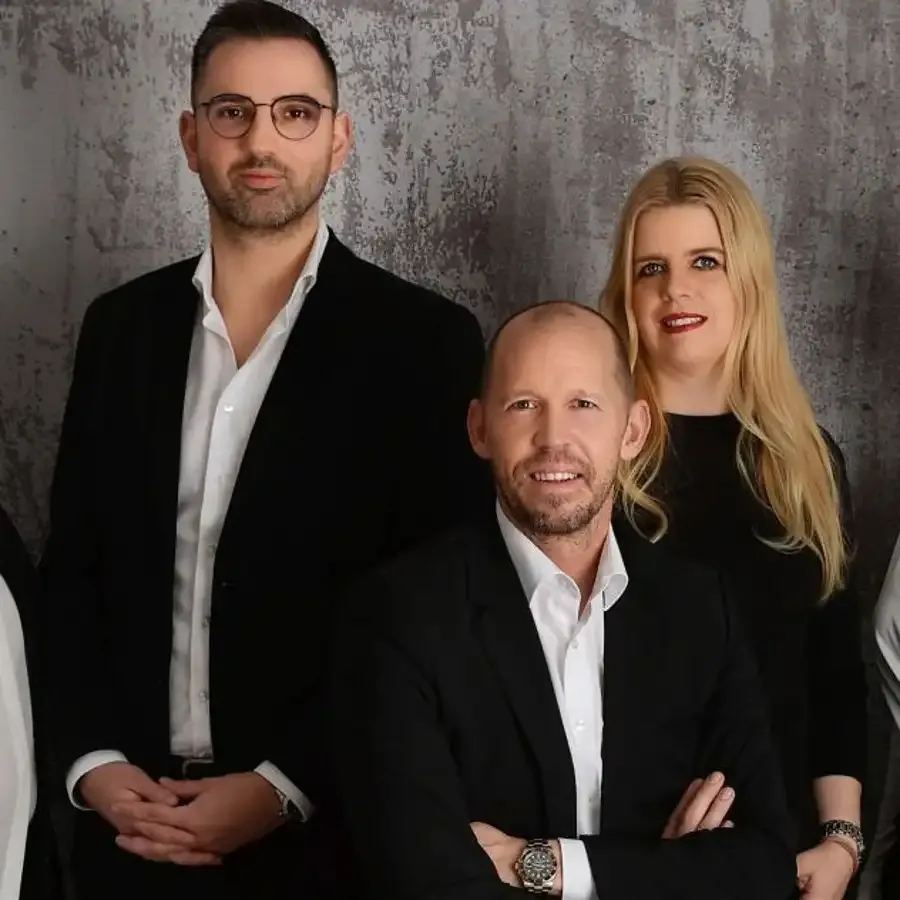 Drei Personen in formeller Kleidung vor einer grauen, strukturierten Wand posieren lächelnd für ein Gruppenfoto.