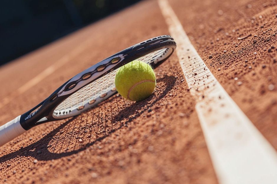 Ein Tennisschläger und ein gelber Tennisball liegen auf einem Sandplatz neben der weißen Linienmarkierung.