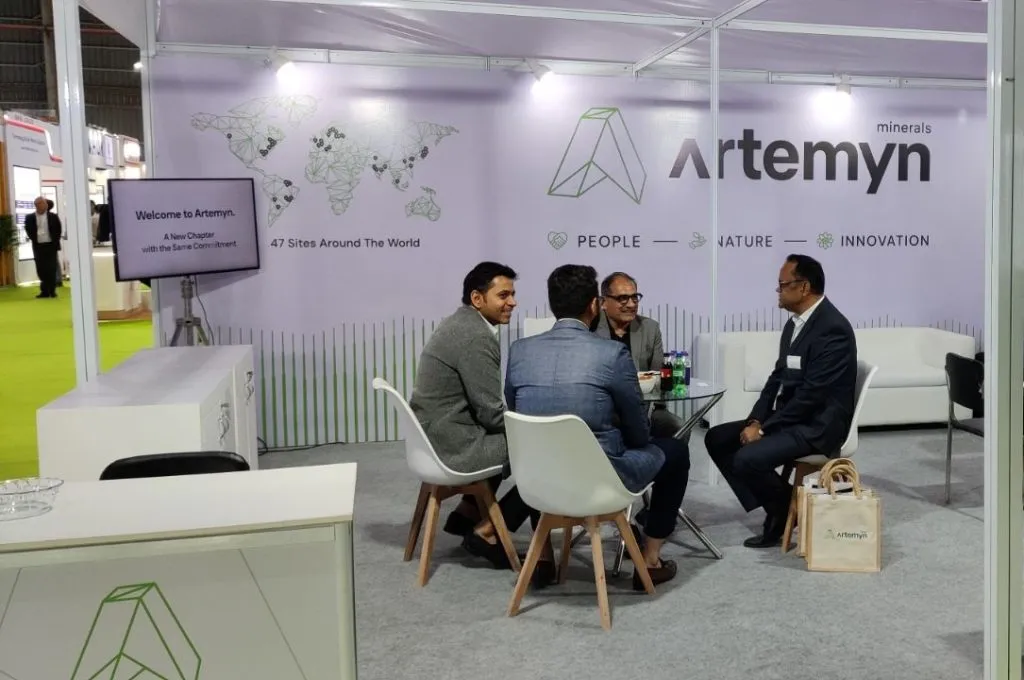 Four men in formal attire sit around a table in a booth, with a backdrop displaying "Artemyn Minerals" and company values.