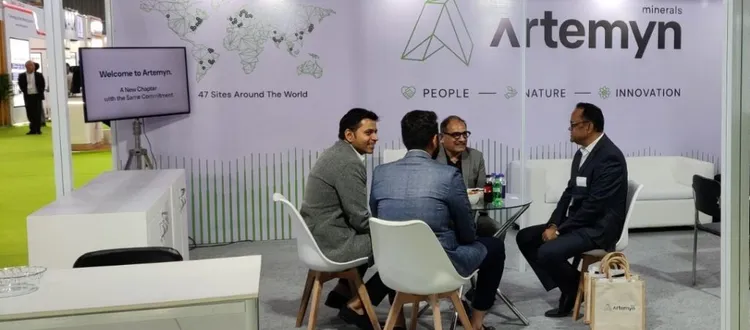 Four men in formal attire sit around a table in a booth, with a backdrop displaying "Artemyn Minerals" and company values.