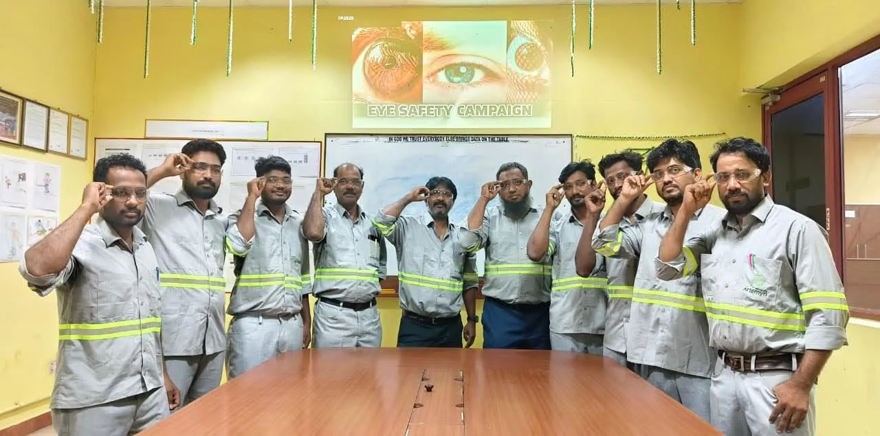 A group of men in safety uniforms participate in an eye safety campaign, each touching their temples, in a room with safety posters.