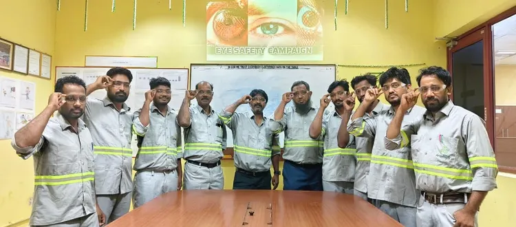 A group of men in safety uniforms participate in an eye safety campaign, each touching their temples, in a room with safety posters.