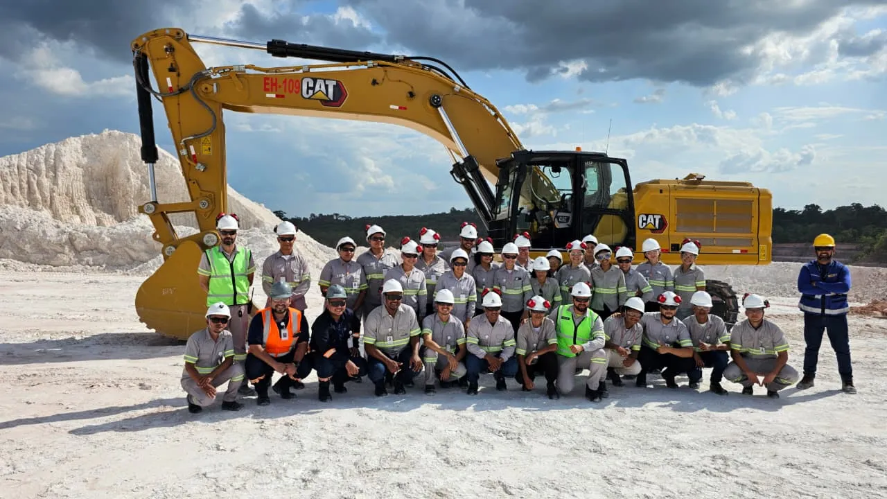 A group of people in safety gear posing in front of a yellow excavator at a construction site with a mound of material in the background.