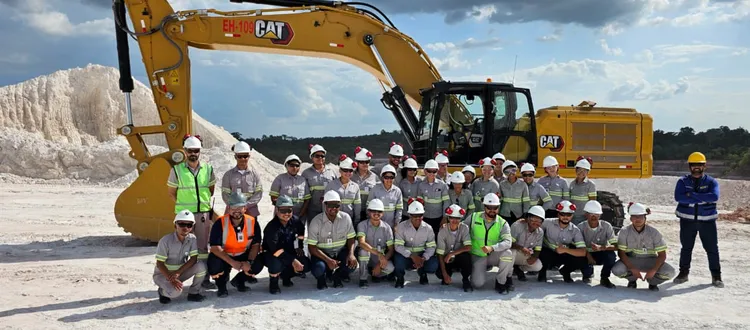 A group of people in safety gear posing in front of a yellow excavator at a construction site with a mound of material in the background.