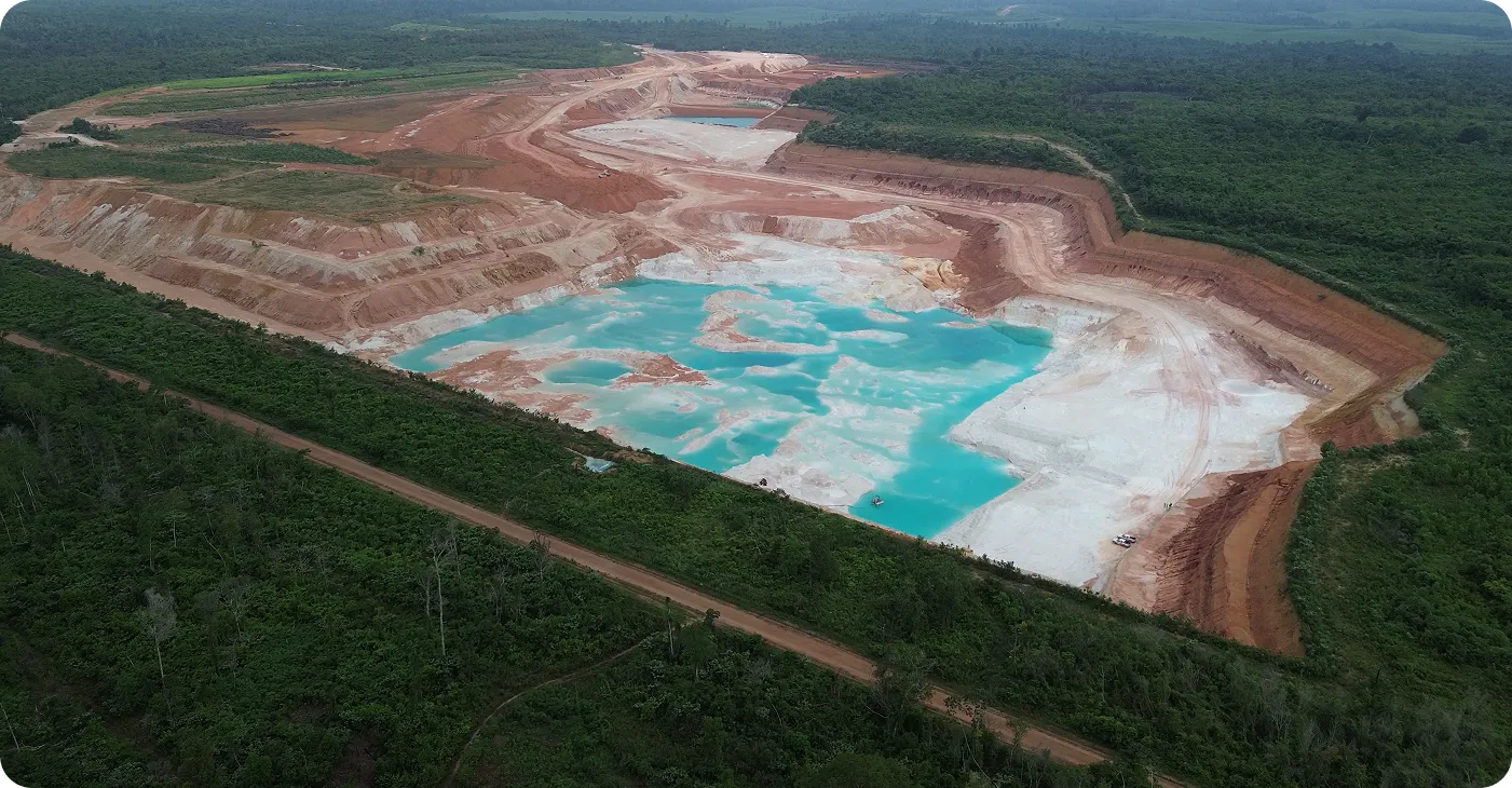 Aerial view of a large open-pit mine with turquoise water pools, surrounded by lush green forest and dirt roads.