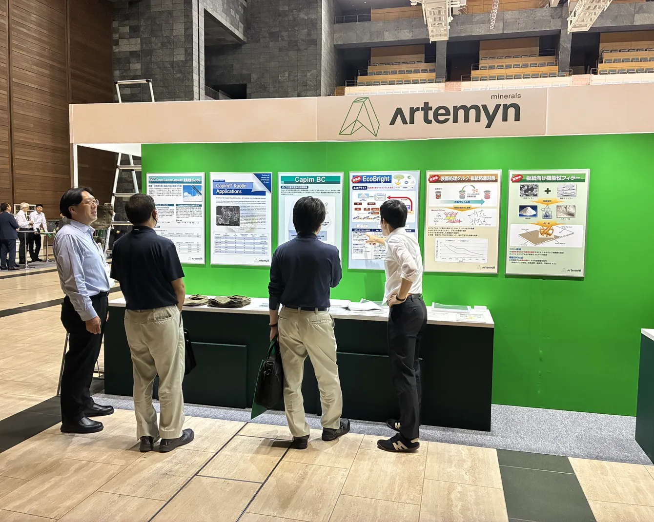 People gather and discuss in front of informational posters at an Artemyn minerals exhibition booth, set against a green backdrop.