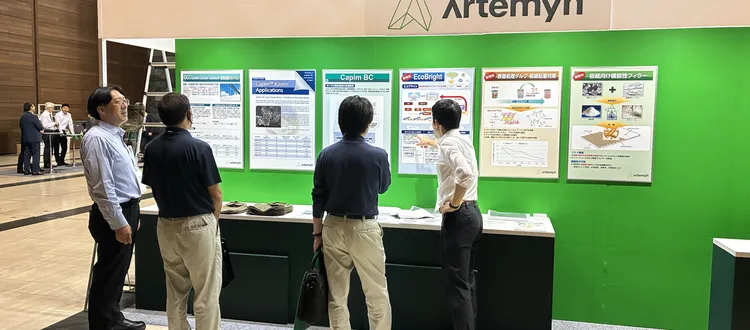 People gather and discuss in front of informational posters at an Artemyn minerals exhibition booth, set against a green backdrop.