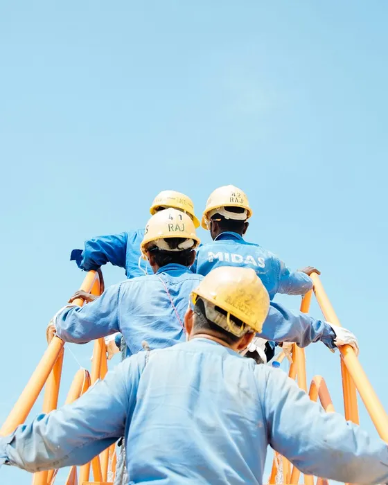 Construction workers in blue uniforms and helmets climb an orange ladder against a clear blue sky.