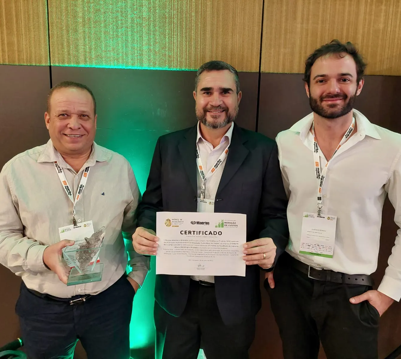 Three men smiling, one holding a certificate and another holding a glass award, wearing lanyards at an indoor event.